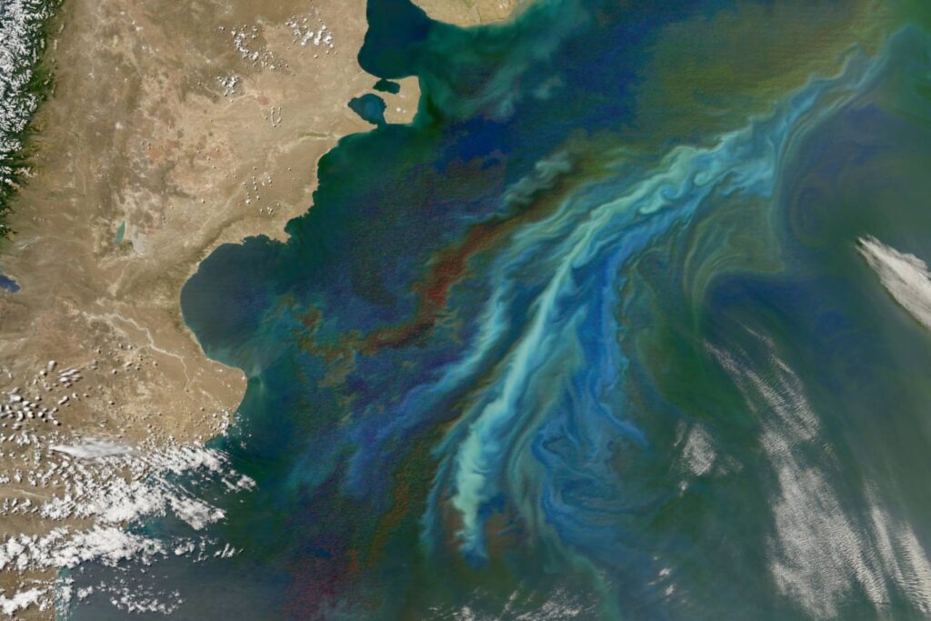 Satellite image showing vivid swirls of blue and green in the ocean near a coast.