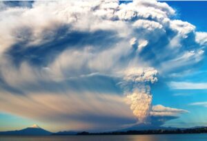 A volcano erupts massively, sending a towering plume of ash into the sky over a calm lake and distant mountains.