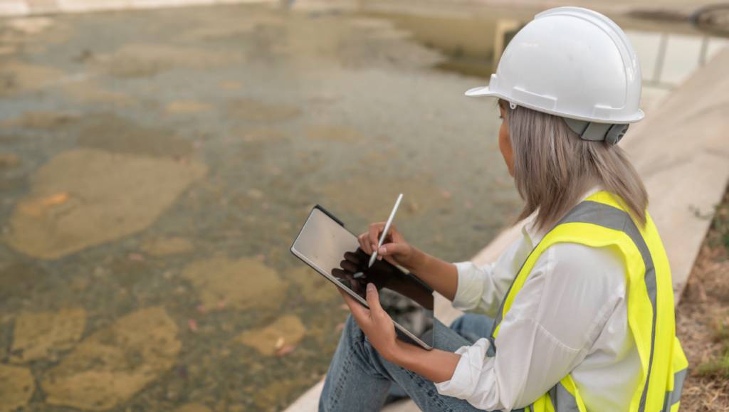 Engineer in hard hat and high-visibility vest using a tablet by a water-filled pit.