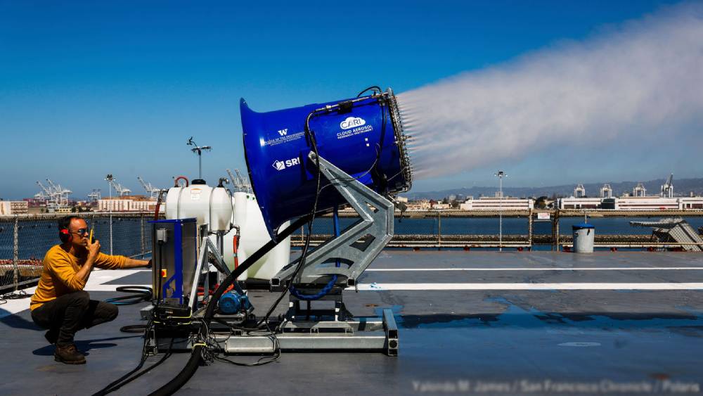 Man tests a large blue snow cannon producing artificial snow on a rooftop, with a city and port in the background.