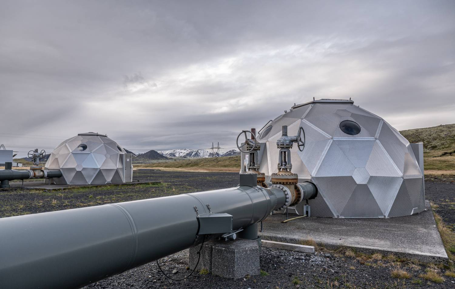Geothermal power plant with hexagonal structures and pipelines in a volcanic landscape, under a cloudy sky.