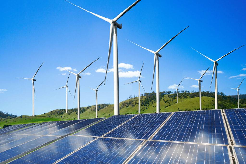 Solar panels in the foreground with wind turbines on a hilly landscape under a clear blue sky.