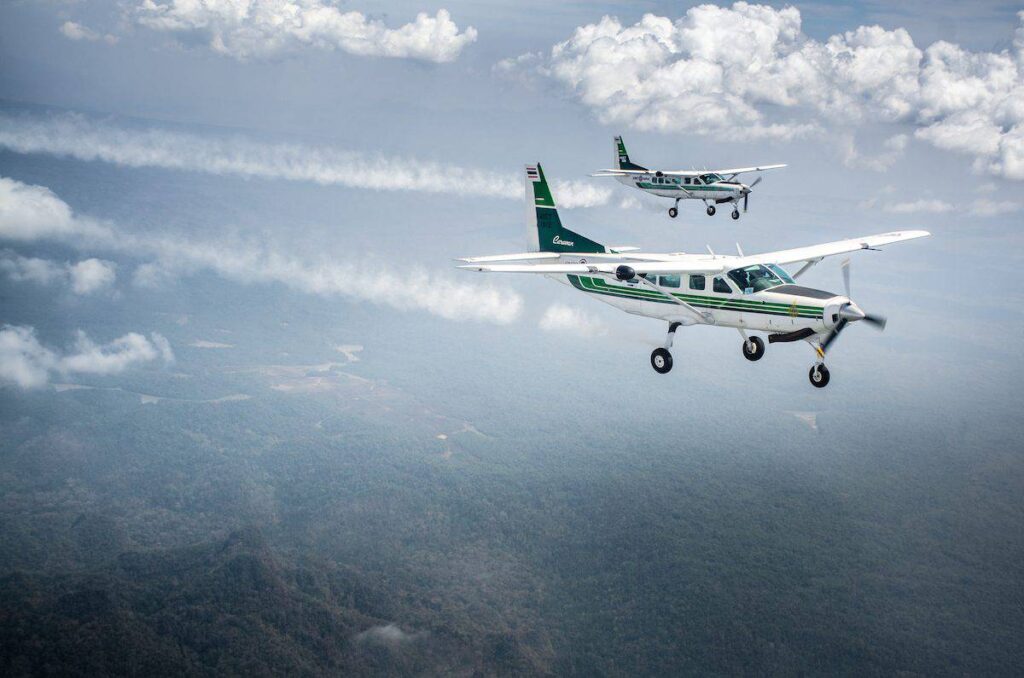 Two light aircraft flying in formation over a lush forest landscape beneath a cloudy sky.