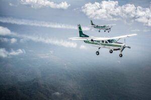 Two light aircraft flying in formation over a lush forest landscape beneath a cloudy sky.