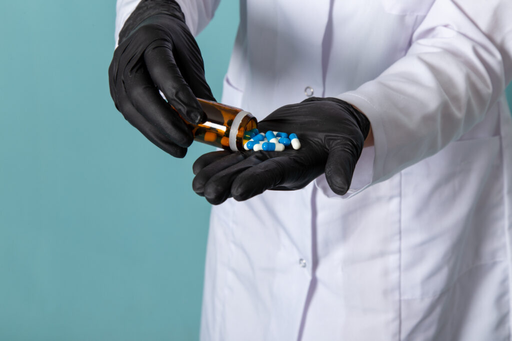 A gloved hand in a lab coat pours blue and white pills from a prescription bottle into the palm of another hand against a teal background.
