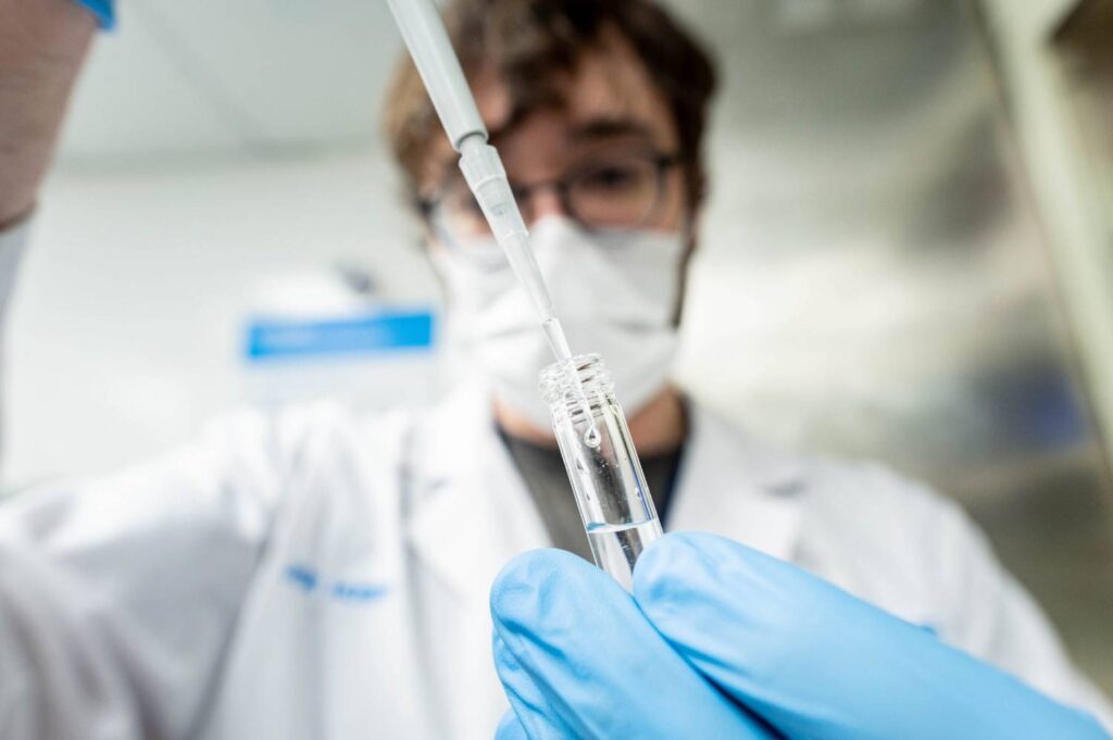 A scientist in a lab coat and mask uses a pipette to handle a liquid sample in a test tube.