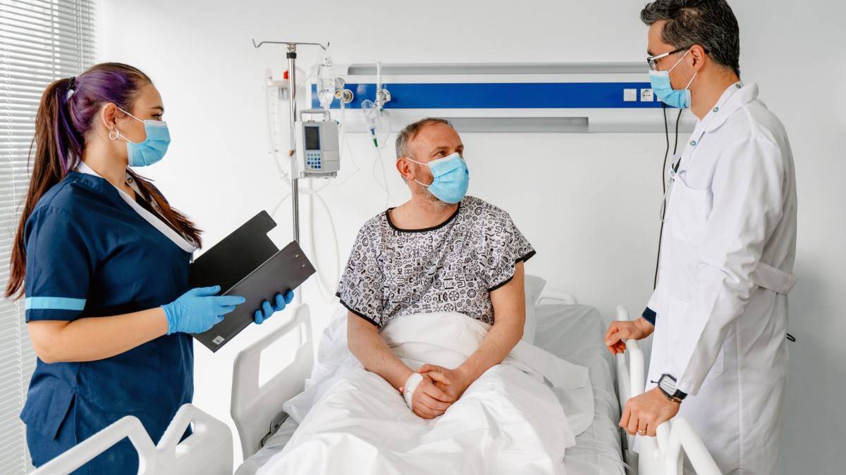 A male patient in hospital bed attended by a nurse and a doctor, all wearing masks.