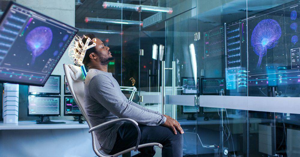 Man wearing a brainwave-reading device sitting in a high-tech lab with multiple screens displaying brain scans.