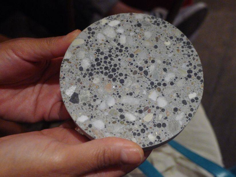 A person holds a round, textured concrete sample featuring a mix of gray tones and various embedded stones and pebbles.