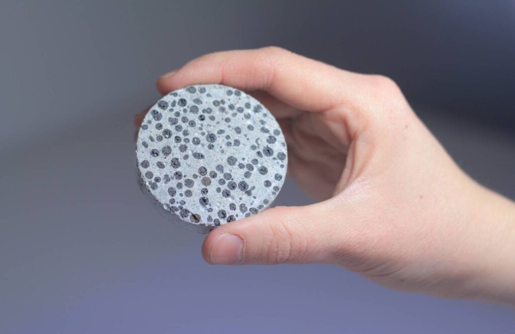 A person holds a circular piece of textured concrete, showcasing various black and gray aggregates against a light background.