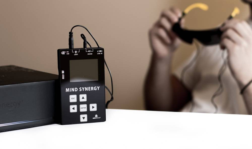 A person holding a headset with a mind synergy device placed beside it on a table.