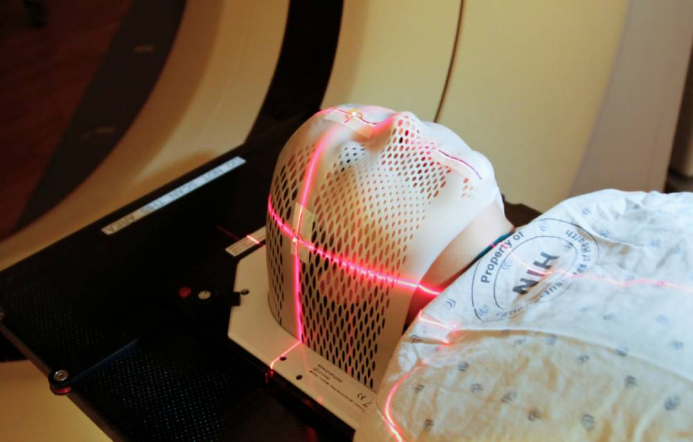 Patient undergoing a medical scan with a head immobilization mask and red laser alignment.
