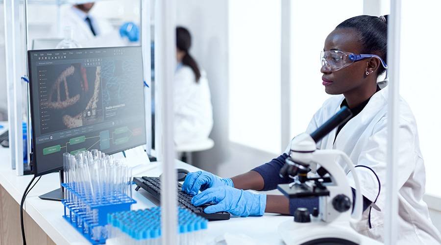 A female scientist using a microscope in a lab, with other researchers and scientific equipment visible in the background.