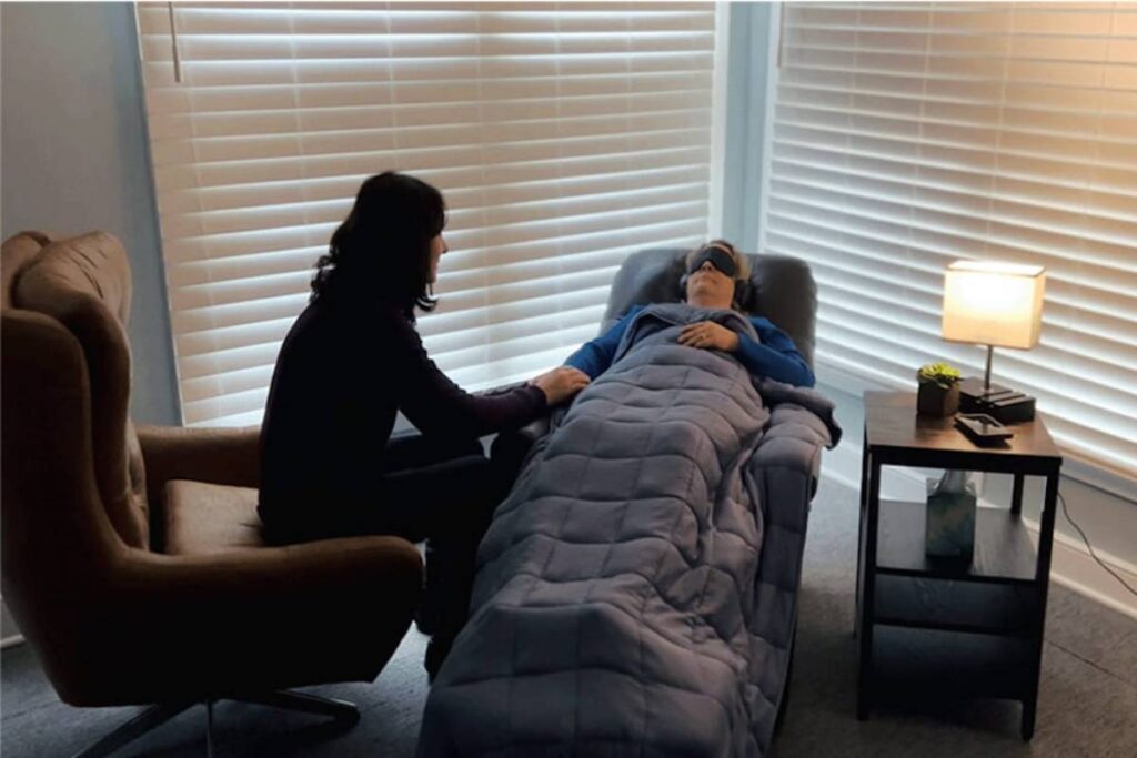A woman in a recliner wearing an eye mask and headphones receives comfort from another woman in a dimly lit room.