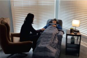 A woman in a recliner wearing an eye mask and headphones receives comfort from another woman in a dimly lit room.