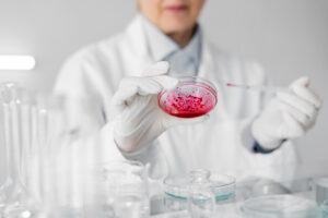 Scientist in lab coat examining a petri dish containing a red substance, surrounded by laboratory glassware.