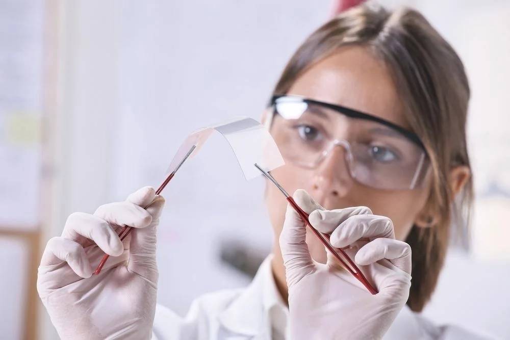 Young female scientist examining a flexible, transparent material in a laboratory, wearing safety goggles and gloves.