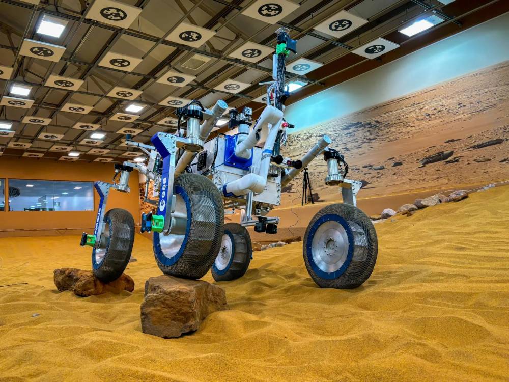 A Mars rover replica on sandy terrain, indoors, simulating a Martian landscape.