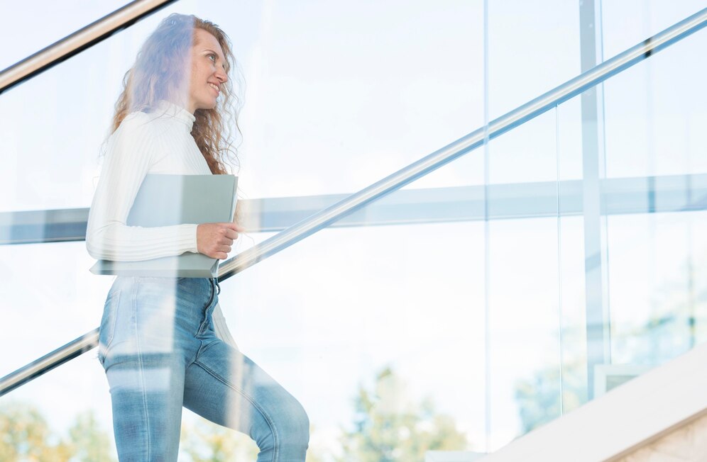 Young woman with curly hair ascending stairs in a modern building while holding a laptop.
