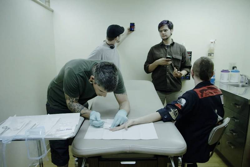 Three men and one woman in a medical room, one man setting up equipment on a bed, another using a smartphone.