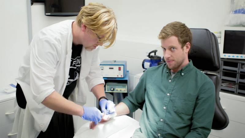 A healthcare professional in a lab coat bandages the hand of a seated male patient in a clinical setting.