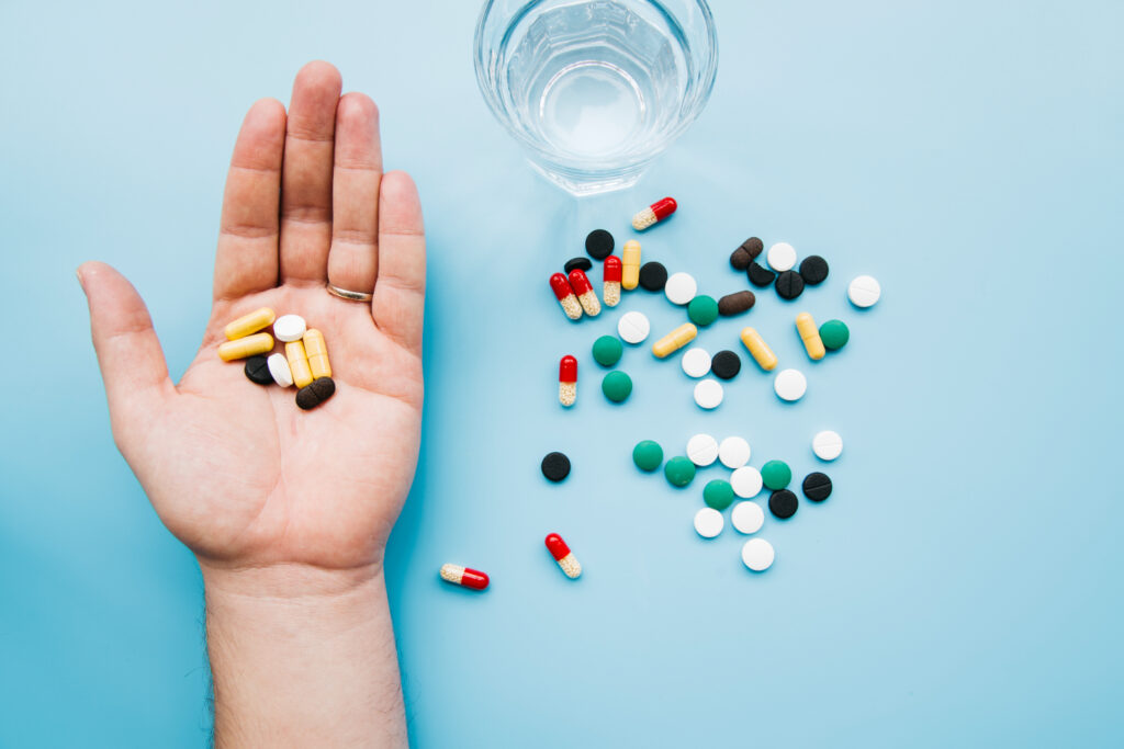 Human hand holding assorted pills, with more capsules and a glass of water on a blue background.