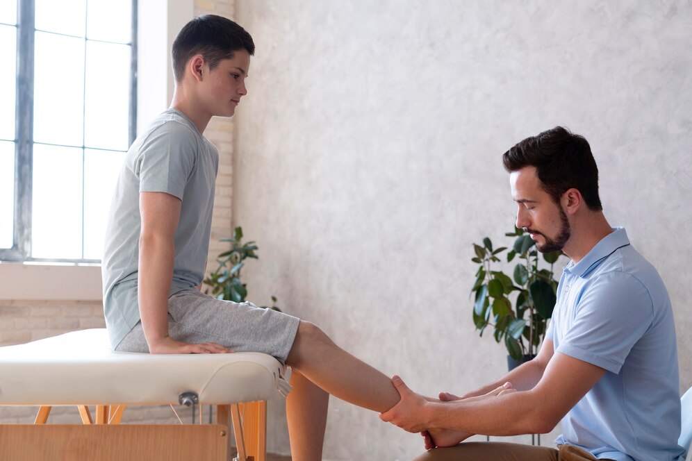 A physical therapist examines a young man's leg in a clinic with a bright window.