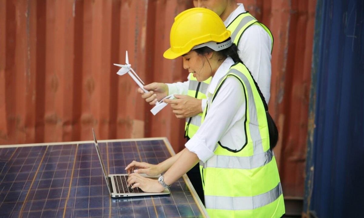 Two engineers in hard hats and reflective vests working with a small wind turbine model and a laptop outdoors.