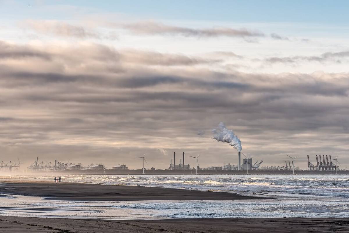 Cloudy beachscape with industrial buildings and smokestacks in the distance, and a few people walking on the shore.
