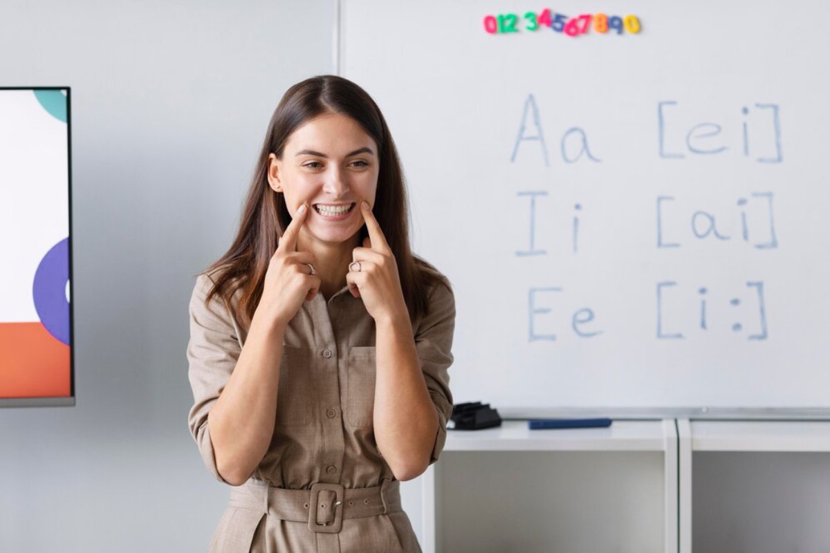 A smiling woman teaching pronunciation, pointing to her mouth, in a classroom with vowel sounds written on a whiteboard behind her.