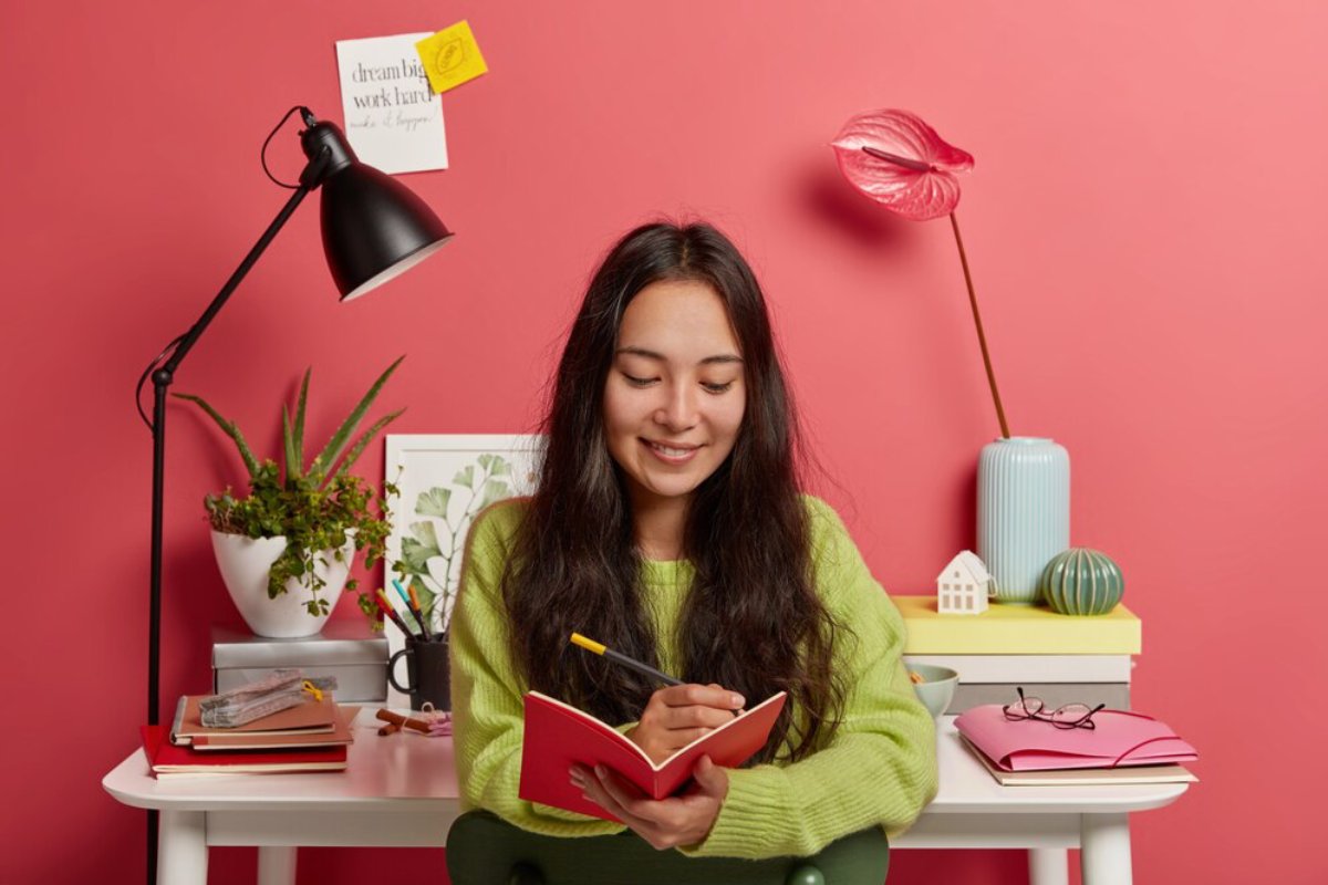 Young woman in a green sweater writing in a red notebook at a colorful, organized desk with plants and decor.
