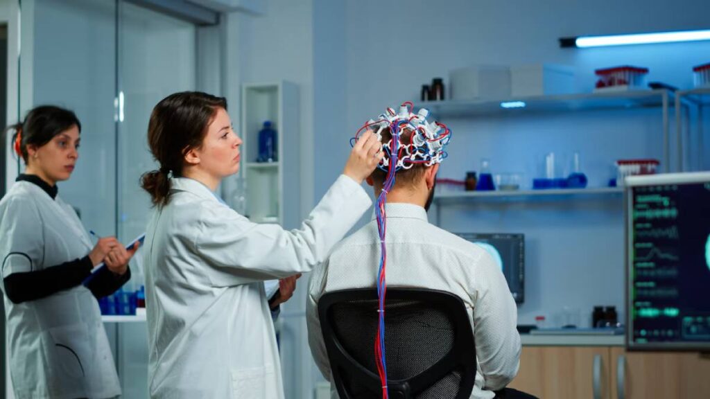 Two female scientists conducting brainwave analysis on a male subject wearing an EEG cap in a laboratory.