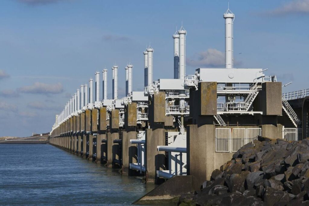 Large storm surge barrier with multiple vertical columns and gates, constructed along a coast under a cloudy sky.