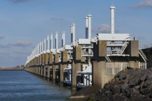 Large storm surge barrier with multiple vertical columns and gates, constructed along a coast under a cloudy sky.