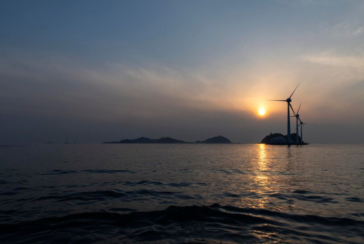 Sunset over calm waters with wind turbines on islands silhouetted against a hazy sky, reflecting golden light on the ocean surface.